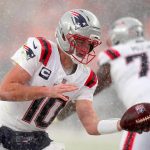 Drake Maye - Ein Footballspieler in der Uniform und dem Helm der New England Patriots mit der Nummer 10 bereitet sich während eines verschneiten Super Bowl LX-Spiels auf die Ballabgabe vor. Es schneit heftig, während die Mannschaftskameraden in weißen Patriots-Uniformen hinter der intensiven Aktion verschwimmen. Diese Beschreibung wurde mit der FootballR KI automatisch generiert.