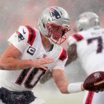 Drake Maye - Ein Footballspieler in der Uniform und dem Helm der New England Patriots mit der Nummer 10 bereitet sich während eines verschneiten Super Bowl LX-Spiels auf die Ballabgabe vor. Es schneit heftig, während die Mannschaftskameraden in weißen Patriots-Uniformen hinter der intensiven Aktion verschwimmen. Diese Beschreibung wurde mit der FootballR KI automatisch generiert.