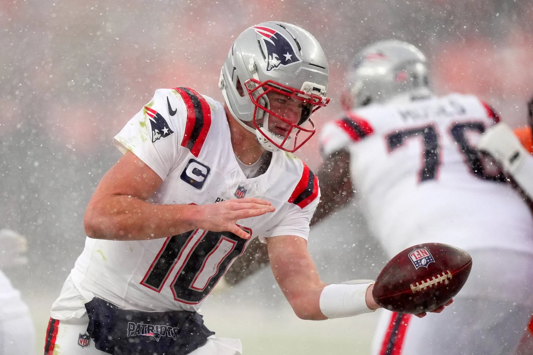 Drake Maye - Ein Footballspieler in der Uniform und dem Helm der New England Patriots mit der Nummer 10 bereitet sich während eines verschneiten Super Bowl LX-Spiels auf die Ballabgabe vor. Es schneit heftig, während die Mannschaftskameraden in weißen Patriots-Uniformen hinter der intensiven Aktion verschwimmen. Diese Beschreibung wurde mit der FootballR KI automatisch generiert.