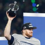 Sam Darnold - Ein Football-Spieler mit einem grauen "CHAMPIONS"-Shirt und einer schwarzen "CONFERENCE CHAMPIONS"-Mütze lächelt und hebt eine Trophäe feierlich in die Höhe. Grünes und blaues Konfetti fällt, als die Seahawks im Sportstadion feiern. Diese Beschreibung wurde mit der FootballR KI automatisch generiert.