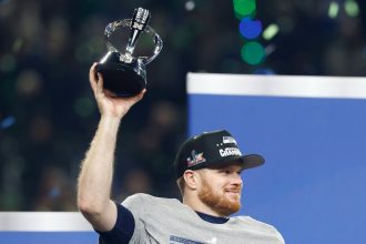 Sam Darnold - Ein Football-Spieler mit einem grauen "CHAMPIONS"-Shirt und einer schwarzen "CONFERENCE CHAMPIONS"-Mütze lächelt und hebt eine Trophäe feierlich in die Höhe. Grünes und blaues Konfetti fällt, als die Seahawks im Sportstadion feiern. Diese Beschreibung wurde mit der FootballR KI automatisch generiert.