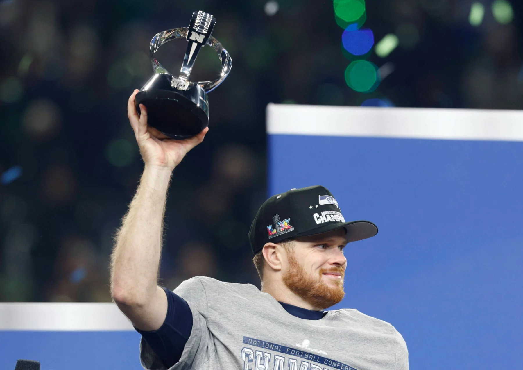Sam Darnold - Ein Football-Spieler mit einem grauen "CHAMPIONS"-Shirt und einer schwarzen "CONFERENCE CHAMPIONS"-Mütze lächelt und hebt eine Trophäe feierlich in die Höhe. Grünes und blaues Konfetti fällt, als die Seahawks im Sportstadion feiern. Diese Beschreibung wurde mit der FootballR KI automatisch generiert.