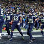 Championship Games - Sechs Footballspieler der Seattle Seahawks in blauen Trikots feiern gemeinsam in der Endzone, einige lächeln und halten die Arme hoch, während die grün-blau gekleidete Menge bei den Championship Games jubelt. Ein Spieler hält einen Football und schaut nach oben. Diese Beschreibung wurde mit der FootballR KI automatisch generiert.