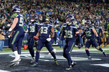 Championship Games - Sechs Footballspieler der Seattle Seahawks in blauen Trikots feiern gemeinsam in der Endzone, einige lächeln und halten die Arme hoch, während die grün-blau gekleidete Menge bei den Championship Games jubelt. Ein Spieler hält einen Football und schaut nach oben. Diese Beschreibung wurde mit der FootballR KI automatisch generiert.