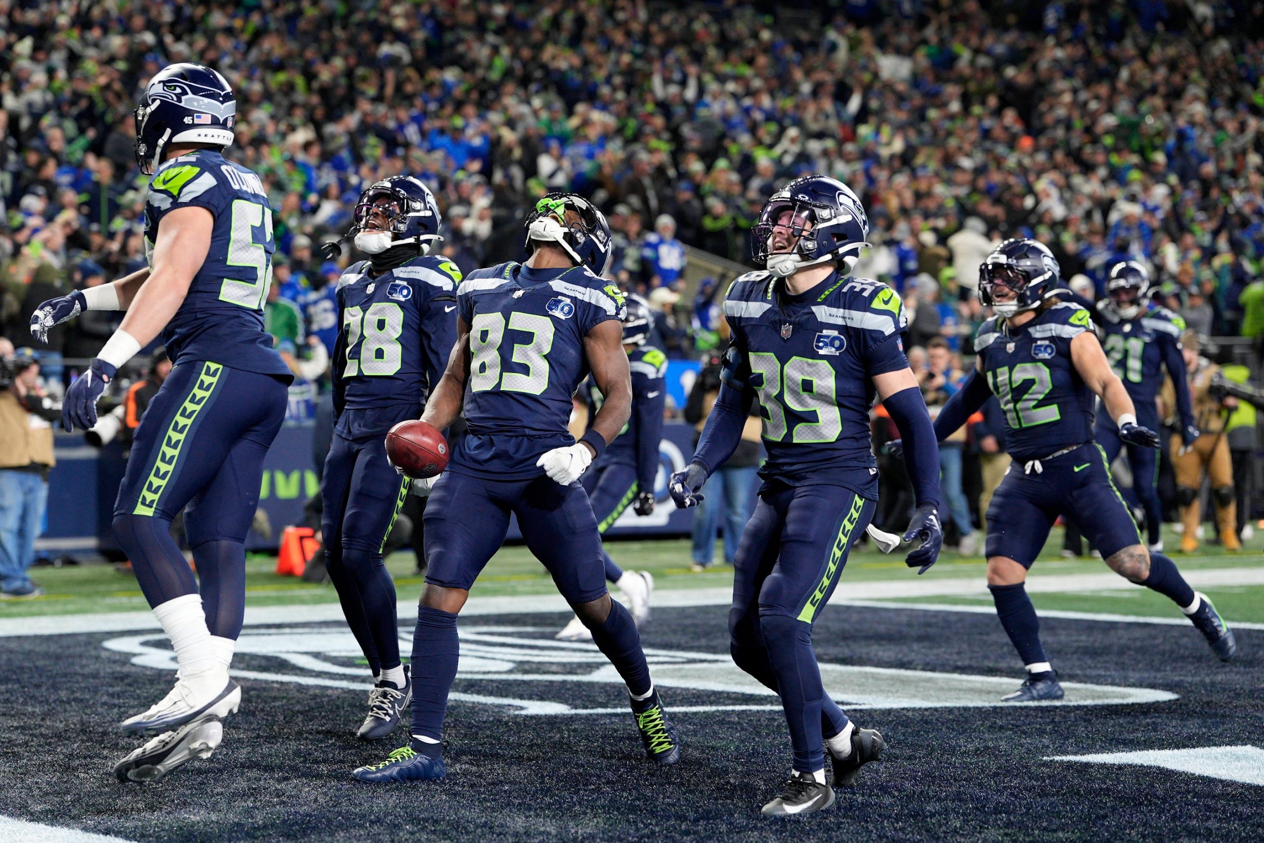 Championship Games - Sechs Footballspieler der Seattle Seahawks in blauen Trikots feiern gemeinsam in der Endzone, einige lächeln und halten die Arme hoch, während die grün-blau gekleidete Menge bei den Championship Games jubelt. Ein Spieler hält einen Football und schaut nach oben. Diese Beschreibung wurde mit der FootballR KI automatisch generiert.