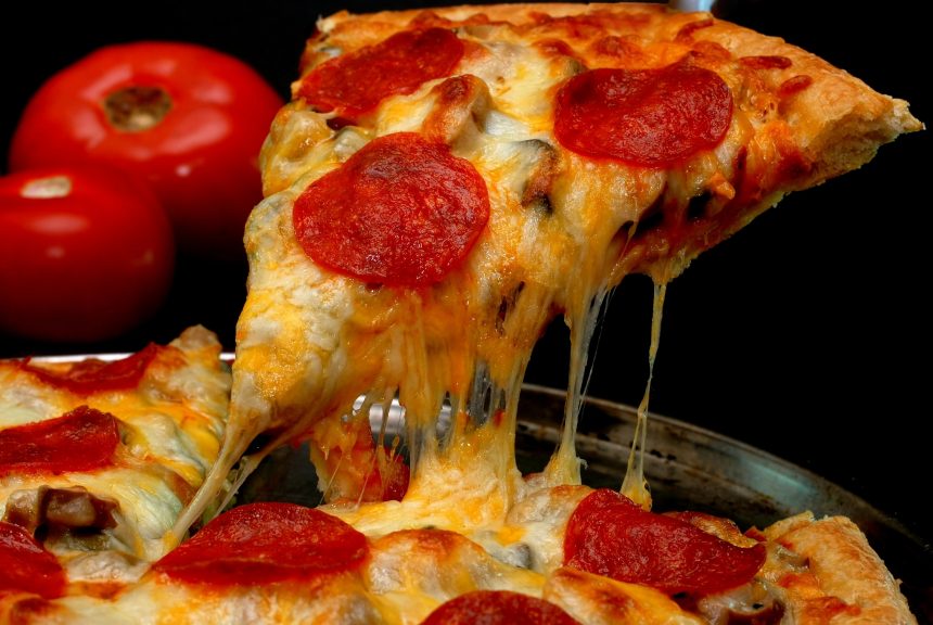 Ein Stück Peperoni-Pizza mit geschmolzenem Käse wird aus einer Pfanne gehoben, im Hintergrund zwei ganze Tomaten - das perfekte Essen für jeden US-Bürger, der den Super Bowl LX schaut. Alt-Text wurde KI-generiert.
