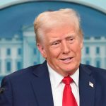 Ein Mann in marineblauem Anzug, weißem Hemd und roter Krawatte steht auf einem Podium vor einem blau-weißen Hintergrund, hinter dem ein Teil eines Emblems zu sehen ist, das an Donald Trump erinnert, der eine Begnadigung für NFL-Profis ankündigt. Alt-Text wurde KI-generiert.