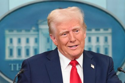 Ein Mann in marineblauem Anzug, weißem Hemd und roter Krawatte steht auf einem Podium vor einem blau-weißen Hintergrund, hinter dem ein Teil eines Emblems zu sehen ist, das an Donald Trump erinnert, der eine Begnadigung für NFL-Profis ankündigt. Alt-Text wurde KI-generiert.