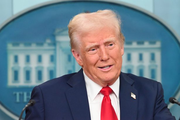 Ein Mann in marineblauem Anzug, weißem Hemd und roter Krawatte steht auf einem Podium vor einem blau-weißen Hintergrund, hinter dem ein Teil eines Emblems zu sehen ist, das an Donald Trump erinnert, der eine Begnadigung für NFL-Profis ankündigt. Alt-Text wurde KI-generiert.