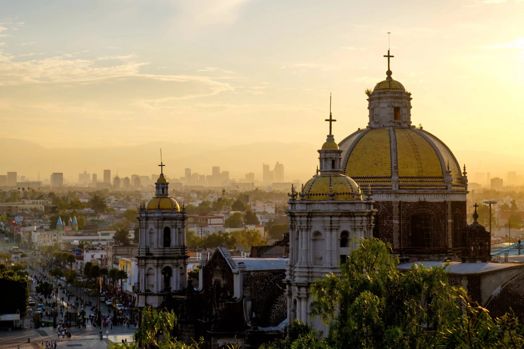 Blick bei Sonnenuntergang auf die Basilika Unserer Lieben Frau von Guadalupe mit ihren Kuppeln und der Skyline von Mexiko Stadt im Hintergrund. Alt-Text wurde KI-generiert.