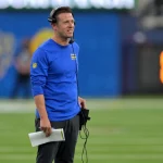 Cheftrainer Mike LaFleur, in einem blauen langärmeligen Hemd und einer schwarzen Hose, steht auf dem Football-Feld der Arizona Cardinals, trägt ein Headset und hält ein Spielblatt in der Hand. Im Hintergrund sind Spieler und ein verschwommenes Stadion zu sehen. Diese Beschreibung wurde mit der FootballR KI automatisch generiert.