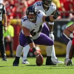 Tyler Linderbaum - Ein Football-Spieler der Offensive Line der Baltimore Ravens in einer weiß-lila Uniform bereitet sich während eines Spiels gegen die Kansas City Chiefs darauf vor, den Ball an der Line of Scrimmage zu spielen. Alt-Text wurde KI-generiert.