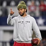 Kyler Murray, der einen grauen "Be Water Bruce Lee"-Kapuzenpulli und eine olivfarbene Mütze trägt, hält auf dem Football-Feld der Cardinals einen Football in der Hand und zeigt nach vorne, um die Energie von Kyler Murray zu kanalisieren. Alt-Text wurde KI-generiert.