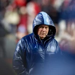 Bill Belichick - Ein Footballtrainer mit blauer Kapuzenjacke und Headset steht während eines Spiels an der Seitenlinie und erinnert an einen Hall of Fame-Führer, der sein Team durch entscheidende Änderungen führt. Alt-Text wurde KI-generiert.