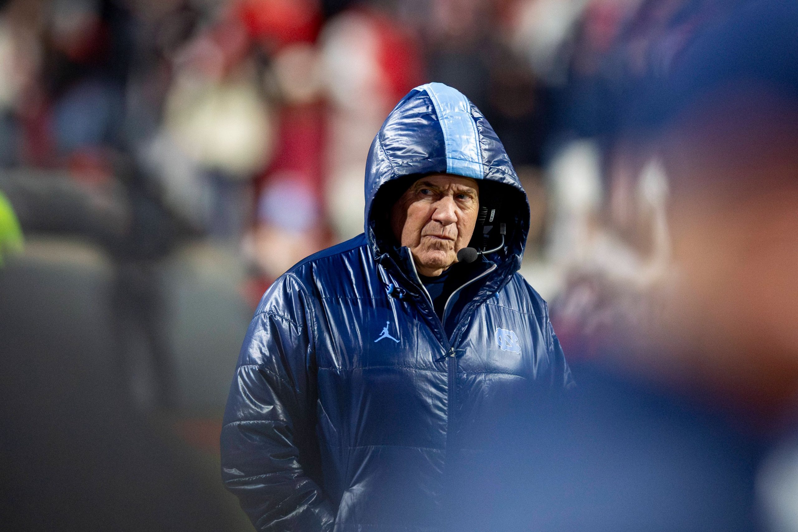 Bill Belichick - Ein Footballtrainer mit blauer Kapuzenjacke und Headset steht während eines Spiels an der Seitenlinie und erinnert an einen Hall of Fame-Führer, der sein Team durch entscheidende Änderungen führt. Alt-Text wurde KI-generiert.