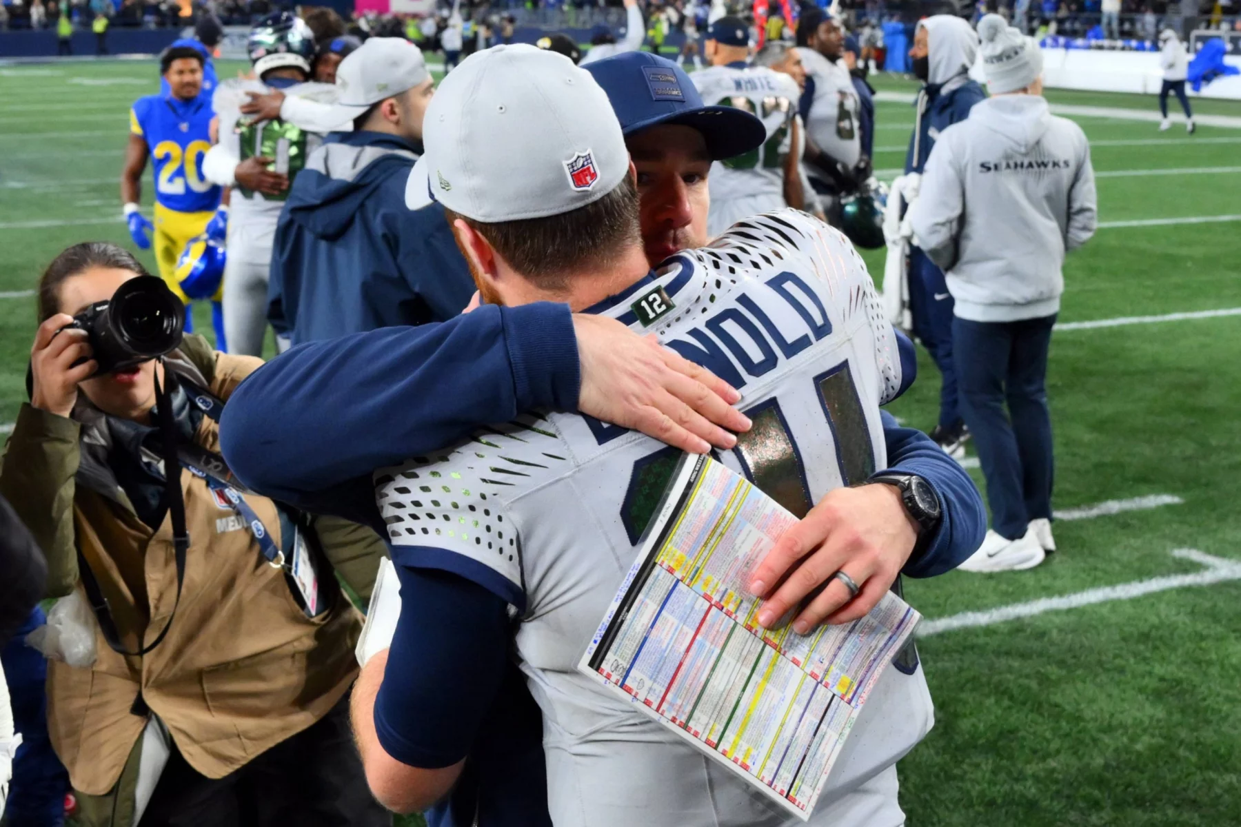 Seattle Seahawks Coach Klint Kubiak - Zwei Footballspieler in Uniform umarmen sich nach einem Spiel auf dem Spielfeld, in der Nähe befindet sich der Cheftrainer der Raiders. Einer hält ein buntes Spielblatt in der Hand. Menschen, darunter auch ein Fotograf, stehen dicht an dicht, während sich die emotionale Szene im Stadion abspielt, mit anderen Spielern im Hintergrund. Diese Beschreibung wurde mit der FootballR KI automatisch generiert.