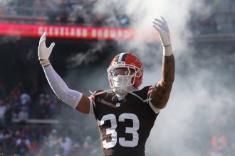 Browns Safety Ronnie Hickman, in voller Uniform, steht mit erhobenen Armen inmitten von Rauch auf einem Stadionfeld, mit Fans im Hintergrund. Alt-Text wurde KI-generiert.