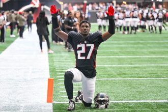 Ein Footballspieler in einem schwarzen Trikot mit der Nummer 27 kniet mit erhobenen Armen auf dem Spielfeld und stellt James Pearce Jr. dar, während Teamkollegen und Mitarbeiter im Hintergrund stehen. Alt-Text wurde KI-generiert.