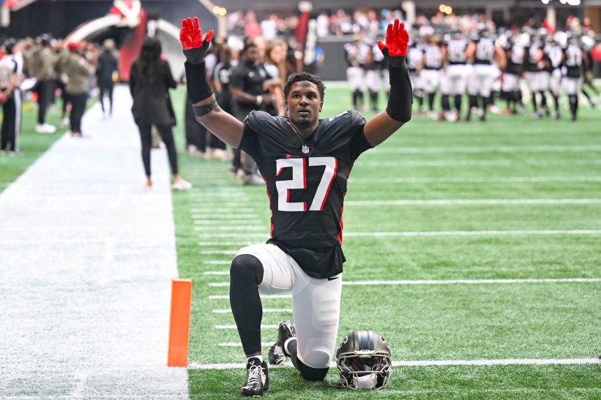 Ein Footballspieler in einem schwarzen Trikot mit der Nummer 27 kniet mit erhobenen Armen auf dem Spielfeld und stellt James Pearce Jr. dar, während Teamkollegen und Mitarbeiter im Hintergrund stehen. Alt-Text wurde KI-generiert.