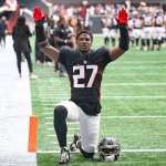 Ein Footballspieler in einem schwarzen Trikot mit der Nummer 27 kniet mit erhobenen Armen auf dem Spielfeld und stellt James Pearce Jr. dar, während Teamkollegen und Mitarbeiter im Hintergrund stehen. Alt-Text wurde KI-generiert.