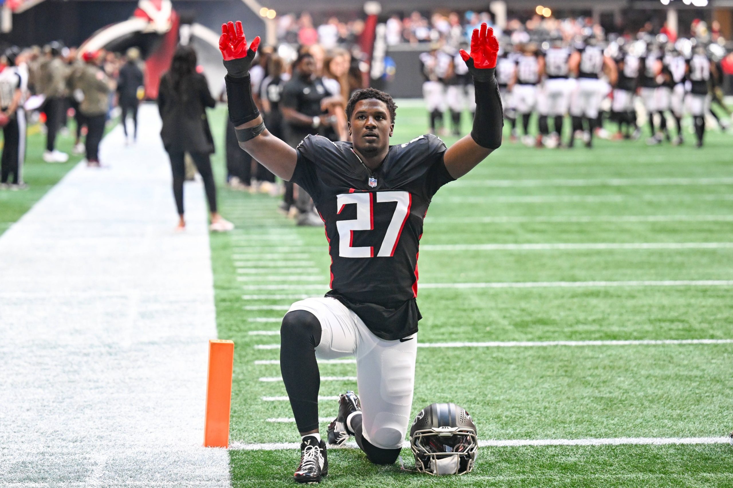 Ein Footballspieler in einem schwarzen Trikot mit der Nummer 27 kniet mit erhobenen Armen auf dem Spielfeld und stellt James Pearce Jr. dar, während Teamkollegen und Mitarbeiter im Hintergrund stehen. Alt-Text wurde KI-generiert.