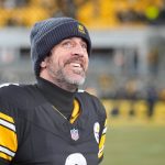 Aaron Rodgers - Ein Football-Spieler in einem Trikot der Pittsburgh Steelers und einer Mütze steht auf dem Spielfeld, blickt nach oben und lächelt, während die Menschenmenge im Hintergrund verschwimmt. Sein hoffnungsvoller Ausdruck erinnert an die Vorfreude auf die Rückkehr eines Stars. Alt-Text wurde KI-generiert.