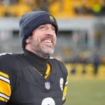 Aaron Rodgers - Ein Football-Spieler in einem Trikot der Pittsburgh Steelers und einer Mütze steht auf dem Spielfeld, blickt nach oben und lächelt, während die Menschenmenge im Hintergrund verschwimmt. Sein hoffnungsvoller Ausdruck erinnert an die Vorfreude auf die Rückkehr eines Stars. Alt-Text wurde KI-generiert.