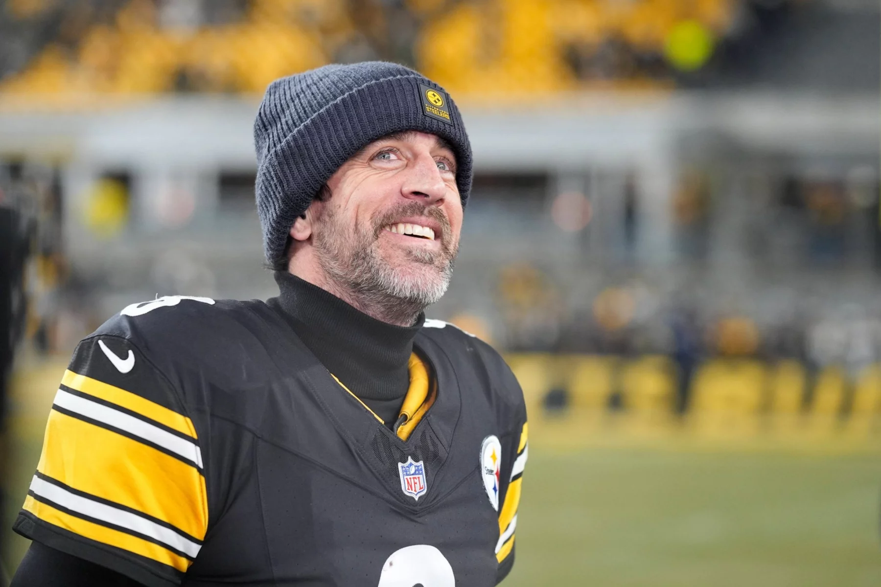 Aaron Rodgers - Ein Football-Spieler in einem Trikot der Pittsburgh Steelers und einer Mütze steht auf dem Spielfeld, blickt nach oben und lächelt, während die Menschenmenge im Hintergrund verschwimmt. Sein hoffnungsvoller Ausdruck erinnert an die Vorfreude auf die Rückkehr eines Stars. Alt-Text wurde KI-generiert.