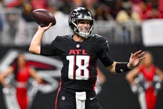 Ein Football-Spieler im Trikot der Atlanta Falcons mit der Nummer 18 bereitet sich darauf vor, während eines Spiels einen Football zu werfen, was die dynamische Energie des Teams auch nach der Verpflichtung von Kirk Cousins unterstreicht. Alt-Text wurde KI-generiert.
