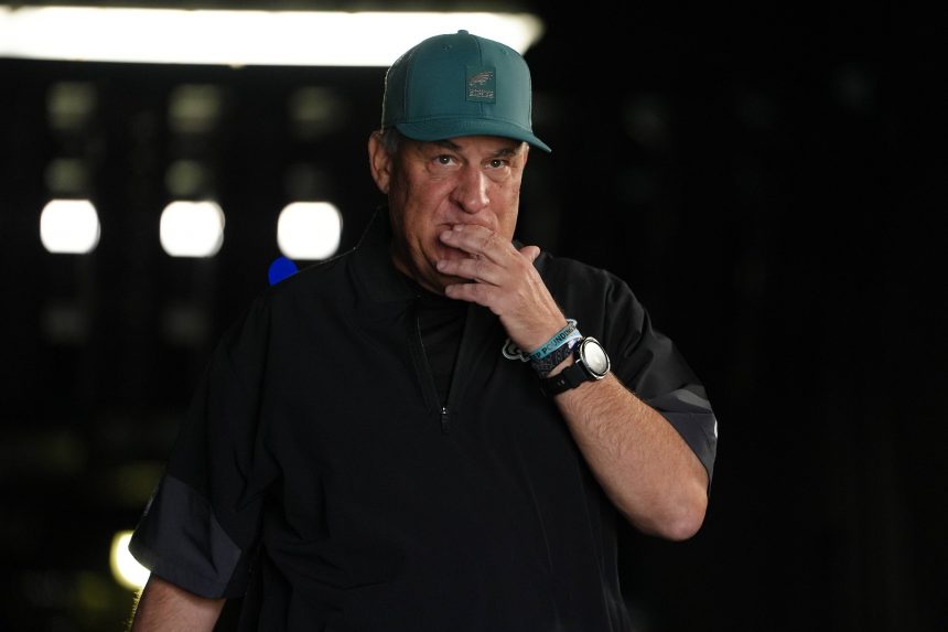 Ein Mann mittleren Alters mit einer blaugrünen Mütze und einer schwarzen Jacke steht in einem schwach beleuchteten Raum und berührt mit der rechten Hand seine Lippen. Er trägt eine Uhr und Armbänder und sieht nachdenklich aus - ähnlich wie Eagles Defensive Coordinator Vic Fangio unter Druck. Helle Lichter verschwimmen hinter ihm. Diese Beschreibung wurde mit der FootballR KI automatisch generiert.