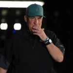 Ein Mann mittleren Alters mit einer blaugrünen Mütze und einer schwarzen Jacke steht in einem schwach beleuchteten Raum und berührt mit der rechten Hand seine Lippen. Er trägt eine Uhr und Armbänder und sieht nachdenklich aus - ähnlich wie Eagles Defensive Coordinator Vic Fangio unter Druck. Helle Lichter verschwimmen hinter ihm. Diese Beschreibung wurde mit der FootballR KI automatisch generiert.