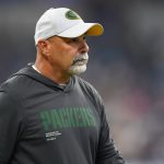 Packers Special Teams Coordinator Rich Bisaccia, in einem Packers-Kapuzenpulli und weißer Mütze, steht während eines Spiels auf dem Spielfeld und schaut zur Seite. Alt-Text wurde KI-generiert.