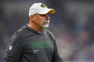 Packers Special Teams Coordinator Rich Bisaccia, in einem Packers-Kapuzenpulli und weißer Mütze, steht während eines Spiels auf dem Spielfeld und schaut zur Seite. Alt-Text wurde KI-generiert.