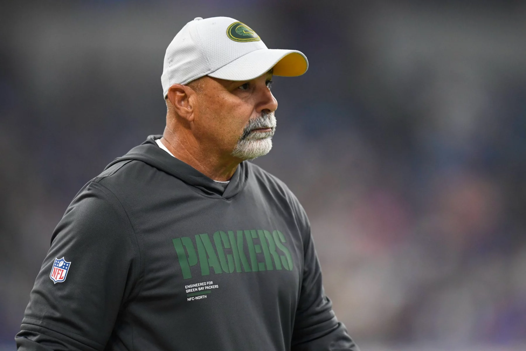 Packers Special Teams Coordinator Rich Bisaccia, in einem Packers-Kapuzenpulli und weißer Mütze, steht während eines Spiels auf dem Spielfeld und schaut zur Seite. Alt-Text wurde KI-generiert.