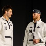Drake Maye und Sam Darnold - Zwei Männer in passenden hellgrauen Pullovern, von denen einer ein Mikrofon in der Hand hält, stehen sich vor einem schlichten dunklen Hintergrund gegenüber und fangen die Aufregung der Eröffnungsfeier des Super Bowl LX ein. Alt-Text wurde KI-generiert.