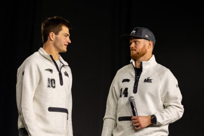 Drake Maye und Sam Darnold - Zwei Männer in passenden hellgrauen Pullovern, von denen einer ein Mikrofon in der Hand hält, stehen sich vor einem schlichten dunklen Hintergrund gegenüber und fangen die Aufregung der Eröffnungsfeier des Super Bowl LX ein. Alt-Text wurde KI-generiert.