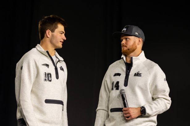 Drake Maye und Sam Darnold - Zwei Männer in passenden hellgrauen Pullovern, von denen einer ein Mikrofon in der Hand hält, stehen sich vor einem schlichten dunklen Hintergrund gegenüber und fangen die Aufregung der Eröffnungsfeier des Super Bowl LX ein. Alt-Text wurde KI-generiert.