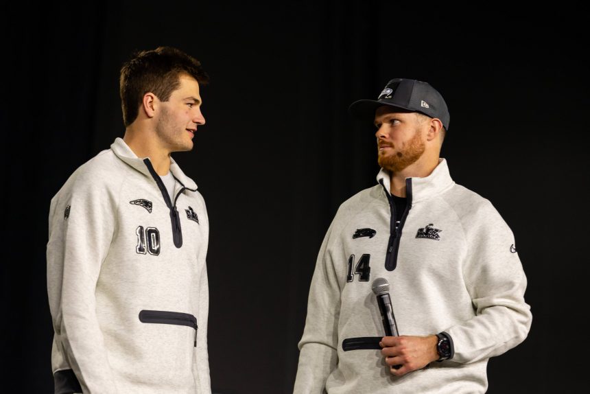 Drake Maye und Sam Darnold - Zwei Männer in passenden hellgrauen Pullovern, von denen einer ein Mikrofon in der Hand hält, stehen sich vor einem schlichten dunklen Hintergrund gegenüber und fangen die Aufregung der Eröffnungsfeier des Super Bowl LX ein. Alt-Text wurde KI-generiert.