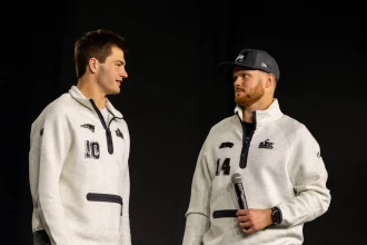 Drake Maye und Sam Darnold - Zwei Männer in passenden hellgrauen Pullovern, von denen einer ein Mikrofon in der Hand hält, stehen sich vor einem schlichten dunklen Hintergrund gegenüber und fangen die Aufregung der Eröffnungsfeier des Super Bowl LX ein. Alt-Text wurde KI-generiert.