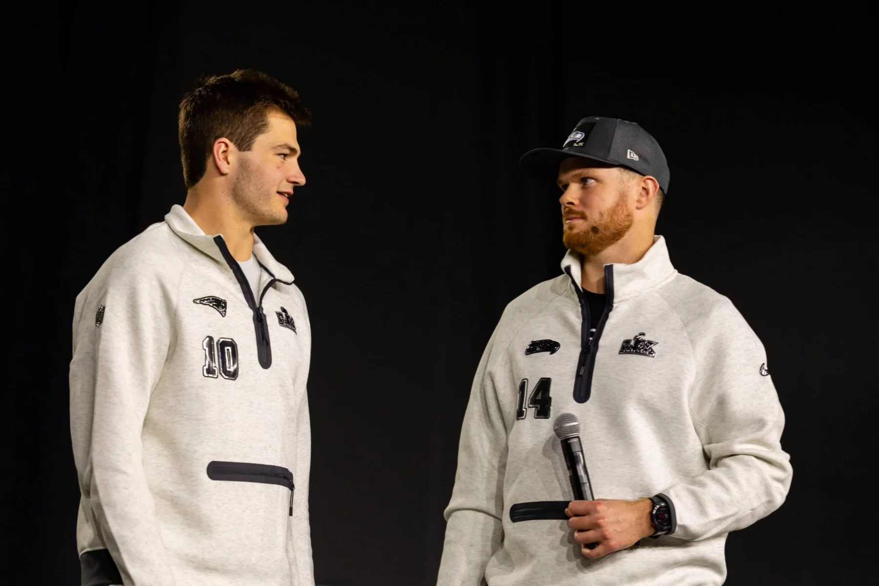 Drake Maye und Sam Darnold - Zwei Männer in passenden hellgrauen Pullovern, von denen einer ein Mikrofon in der Hand hält, stehen sich vor einem schlichten dunklen Hintergrund gegenüber und fangen die Aufregung der Eröffnungsfeier des Super Bowl LX ein. Alt-Text wurde KI-generiert.
