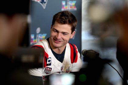 Drake Maye - Ein Footballspieler im Trikot der New England Patriots spricht auf einer Pressekonferenz, lächelnd, mit Mikrofonen und Kameras vor sich, über die Super Bowl-Ambitionen des Teams. Alt-Text wurde KI-generiert.