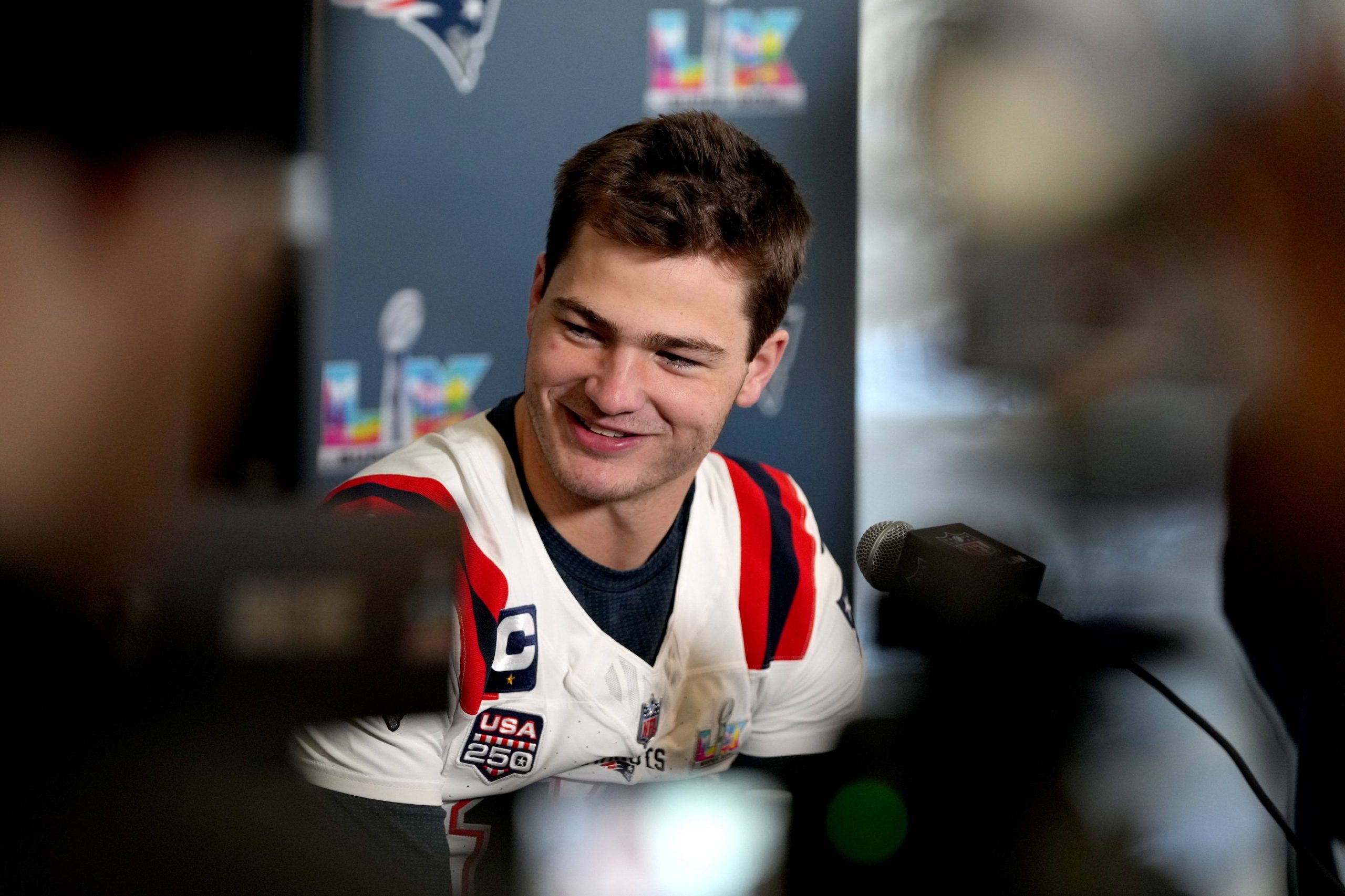 Drake Maye - Ein Footballspieler im Trikot der New England Patriots spricht auf einer Pressekonferenz, lächelnd, mit Mikrofonen und Kameras vor sich, über die Super Bowl-Ambitionen des Teams. Alt-Text wurde KI-generiert.