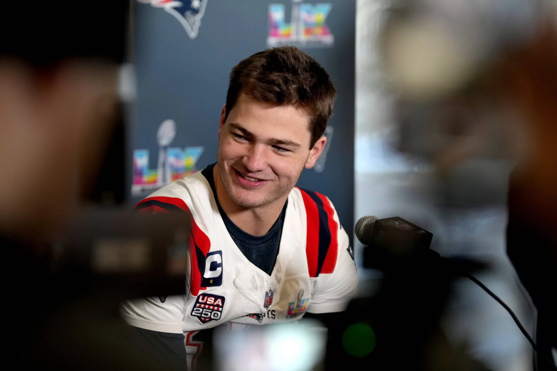 Drake Maye - Ein Footballspieler im Trikot der New England Patriots spricht auf einer Pressekonferenz, lächelnd, mit Mikrofonen und Kameras vor sich, über die Super Bowl-Ambitionen des Teams. Alt-Text wurde KI-generiert.