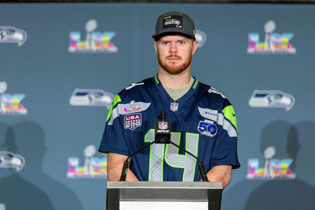 Sam Darnold - Ein Footballspieler in einem Seattle Seahawks-Trikot und einer Kappe steht während einer Presseveranstaltung auf einem Podium, mit Super Bowl- und NFL-Logos im Hintergrund, und spricht über Sam Darnolds jüngste Verletzung. Alt-Text wurde KI-generiert.