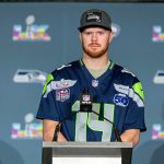 Sam Darnold - Ein Footballspieler in einem Seattle Seahawks-Trikot und einer Kappe steht während einer Presseveranstaltung auf einem Podium, mit Super Bowl- und NFL-Logos im Hintergrund, und spricht über Sam Darnolds jüngste Verletzung. Alt-Text wurde KI-generiert.