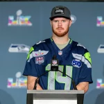 Sam Darnold - Ein Footballspieler in einem Seattle Seahawks-Trikot und einer Kappe steht während einer Presseveranstaltung auf einem Podium, mit Super Bowl- und NFL-Logos im Hintergrund, und spricht über Sam Darnolds jüngste Verletzung. Alt-Text wurde KI-generiert.