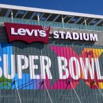Super Bowl LX - Außenansicht des Levi's Stadium mit großem "SUPER BOWL 60"-Schild und Arbeitern an der Glasfassade unter strahlend blauem Himmel. Alt-Text wurde KI-generiert.