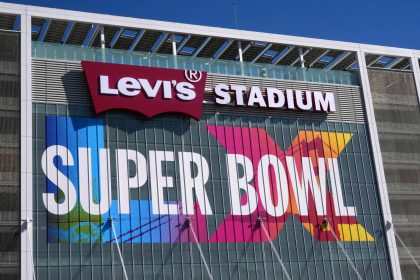 Super Bowl LX - Außenansicht des Levi's Stadium mit großem "SUPER BOWL 60"-Schild und Arbeitern an der Glasfassade unter strahlend blauem Himmel. Alt-Text wurde KI-generiert.