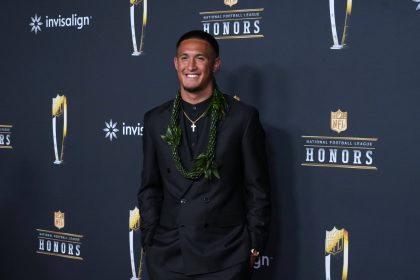 Tetairoa McMillan - Ein Mann in einem schwarzen Anzug mit einem grünen Blumenkranz lächelt auf dem roten Teppich einer NFL-Ehrungsveranstaltung, bei der der Offensive Rookie of the Year geehrt wird, während im Hintergrund die Logos der Veranstaltung und die Banner der Sponsoren zu sehen sind. Alt-Text wurde KI-generiert.