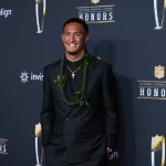 Tetairoa McMillan - Ein Mann in einem schwarzen Anzug mit einem grünen Blumenkranz lächelt auf dem roten Teppich einer NFL-Ehrungsveranstaltung, bei der der Offensive Rookie of the Year geehrt wird, während im Hintergrund die Logos der Veranstaltung und die Banner der Sponsoren zu sehen sind. Alt-Text wurde KI-generiert.